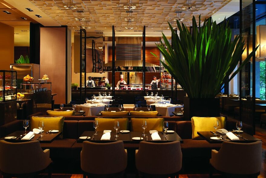 Interior of Collins Kitchen showing dark wooden tables, soft upholstered chairs and the open kitchen in the background.