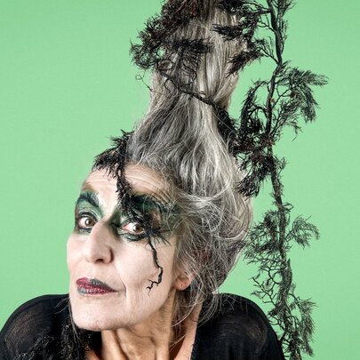 A woman with theatrical witch makeup and an elaborate, tall gray hairstyle intertwined with dark green foliage poses against a solid light green background.
