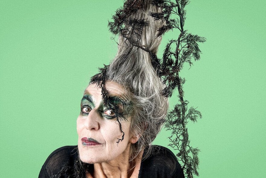 A woman with theatrical witch makeup and an elaborate, tall gray hairstyle intertwined with dark green foliage poses against a solid light green background.