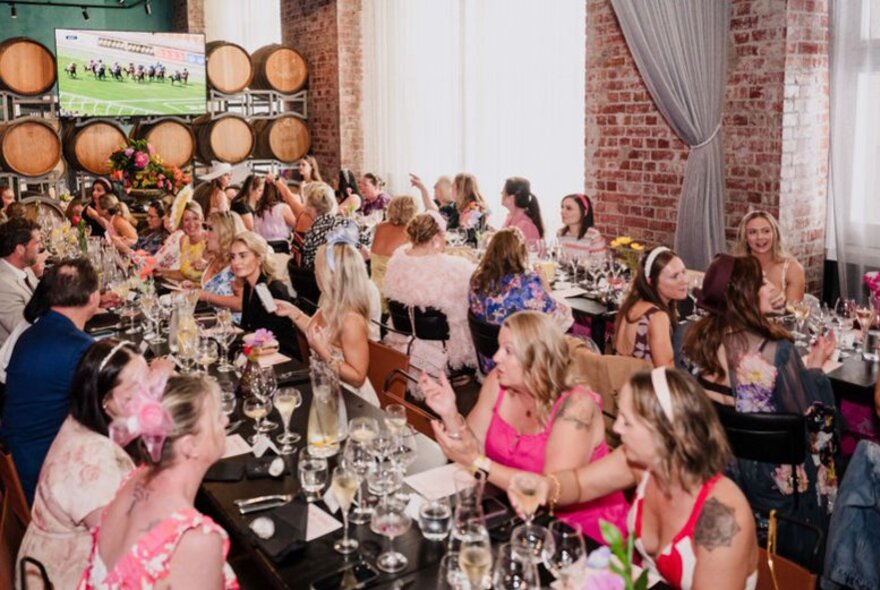 Groups of people dressed in spring racing finery enjoying a meal in a restaurant with live racing on the screen behind them.