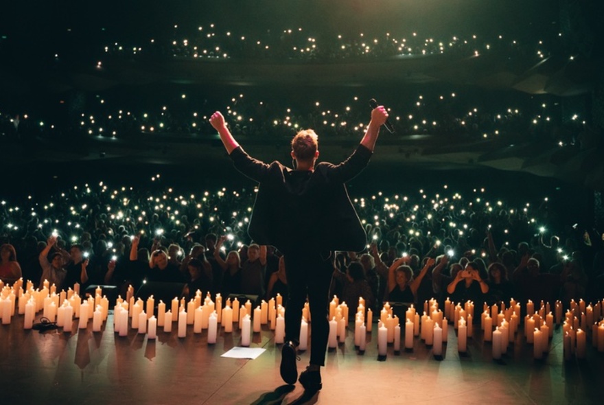 A man on a candlelit stage with outstretched arms, seen from behind with the audience in front of him.