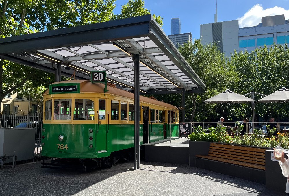 A yellow and green retro Melbourne tram parked under a roof with people sitting outside under umbrellas.