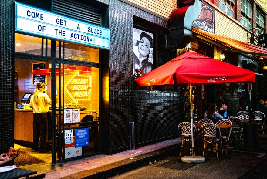 A small restaurant with neon lights and seating outside under a large red umbrella.