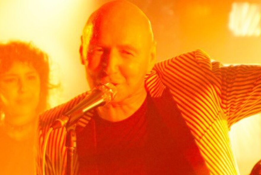 Man with no hair singing into a microphone live on stage, another person on stage in the background, the entire image bathed in a warm yellow-orange light.