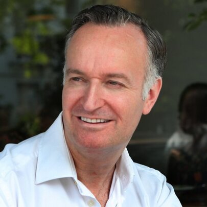 Scottish author Andrew O'Hagan, wearing a white shirt, seated in a dark room with leafy background.