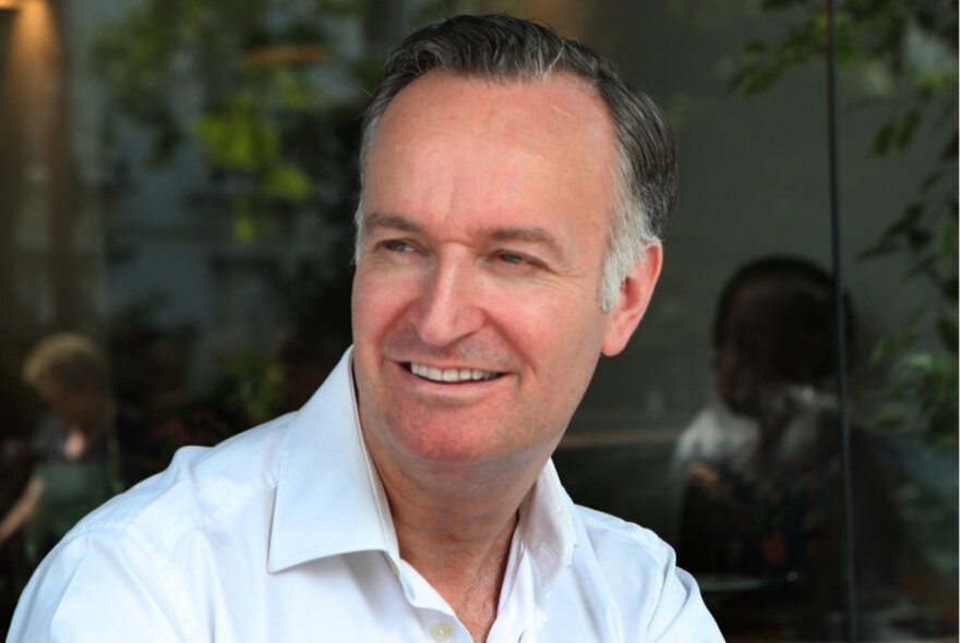 Scottish author Andrew O'Hagan, wearing a white shirt, seated in a dark room with leafy background.