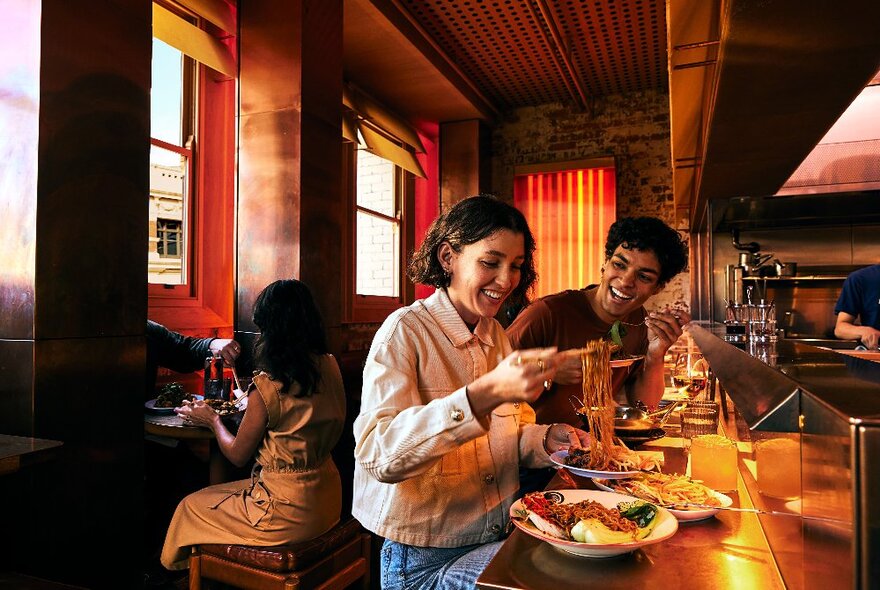 Two people dining at a bar on counter stools in a moodily lit dining area, on dishes including noodles and chips.