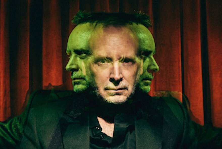 A spooky image of a man in front of a red curtain, with green transparent left and right profiles juxtaposed on his face.