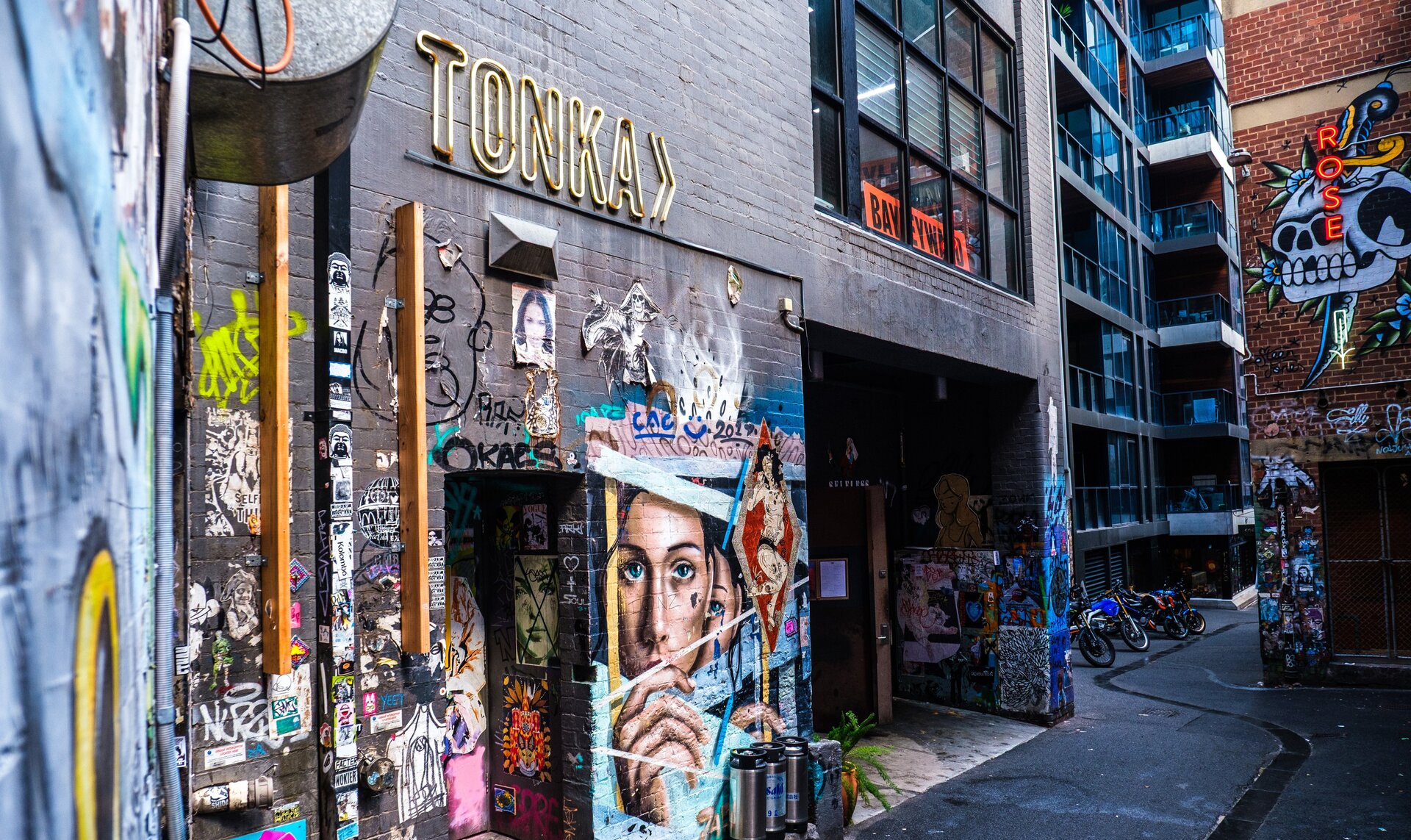 A black brick wall in an alleyway with street art and a sign on it that says Tonka.