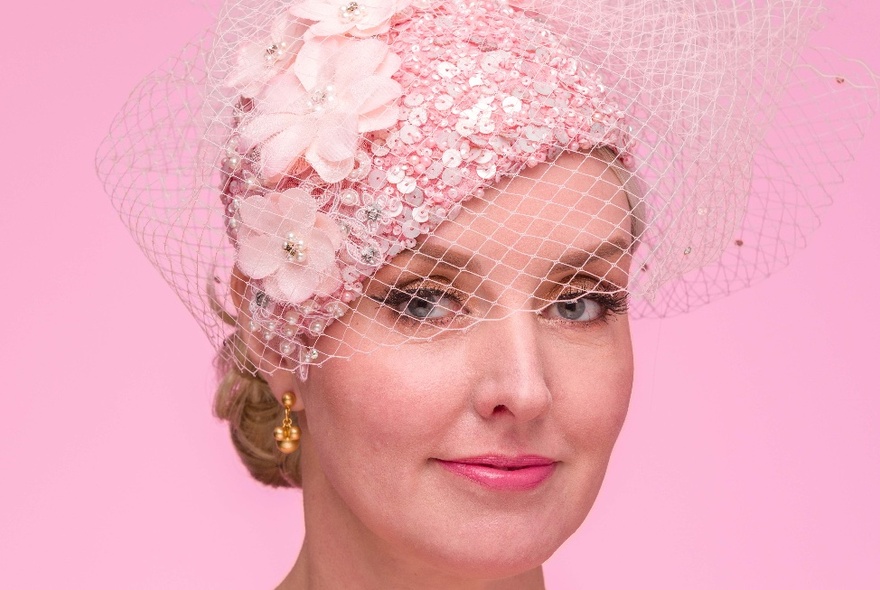 A headshot of a woman wearing a pink fascinator with netting over half her face.