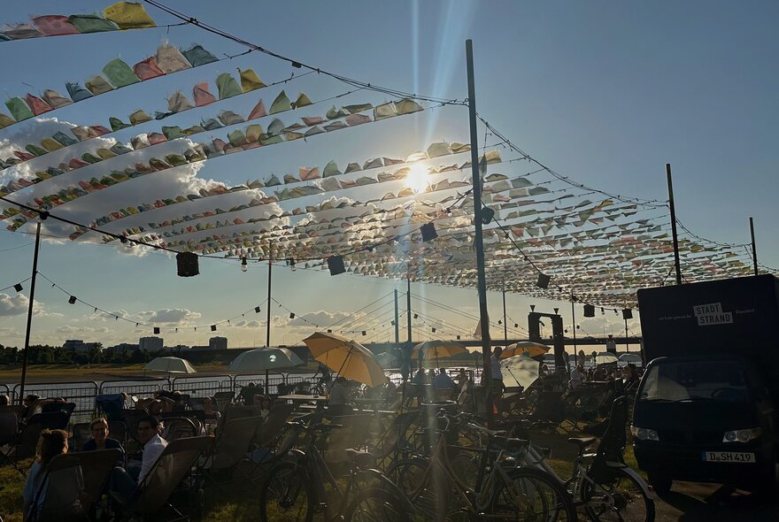 People enjoying themselves outdoors near a riverside, some under shade umbrellas, with colourful bunting strung up above them flapping against the blue sky background.
