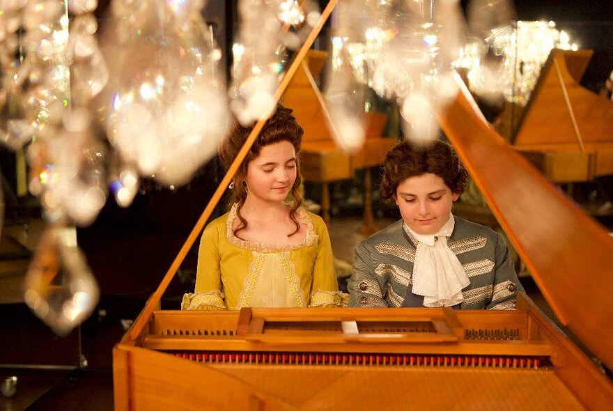 Two teenagers wearing 18th century clothing sitting playing a piano, with chandeliers hanging in the foreground.