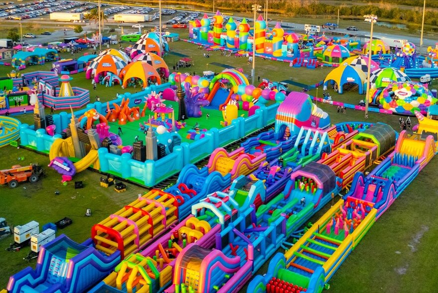Aerial view of large colourful inflatables including obstacle courses and jumping castles in a large flat grass area.