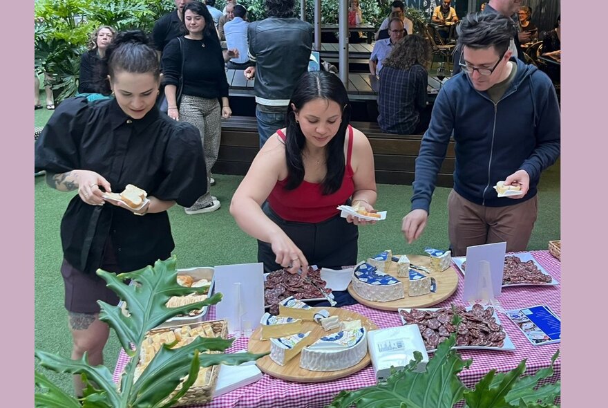 Three people helping themselves to cheese and snacks from a table in an outdoor terrace setting.