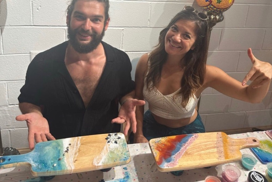 Two people showing off their resin-decorated breadboards in a workshop setting.