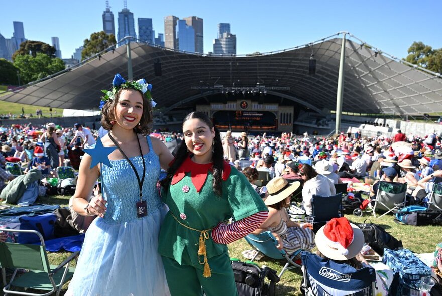 Performers dressed as a fairy and elf standing in sunshine in front of crowds gathered at Myer Music Bowl.