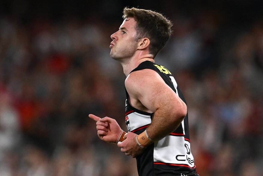 A St Kilda AFL player looking pleased with his forearms close to his side, his finger pointing,  and a blurred stadium crowd behind him.