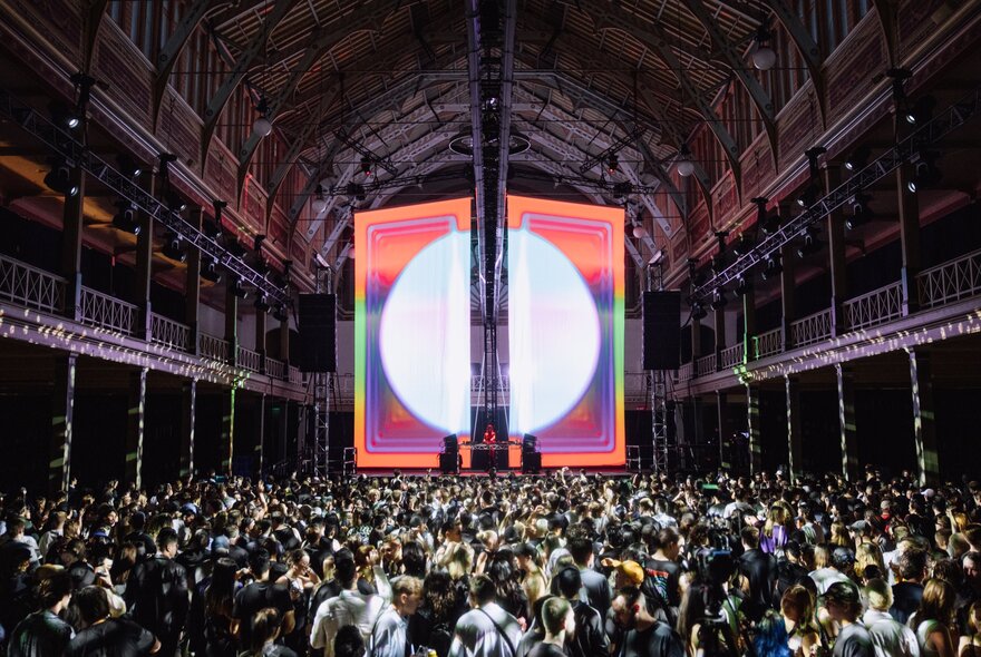 A crowd of festival attendees dancing inside the dim interior of the Royal Exhibition Building, to a DJ playing music on the stage, against a large backdrop of a white circle. 