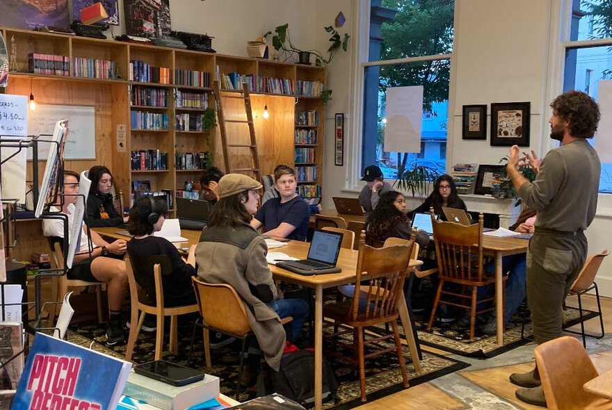 Teenagers sitting and working around two large tables in a room, some with laptops open in front of them, bookshelves in the background and a standing teacher talking to them.