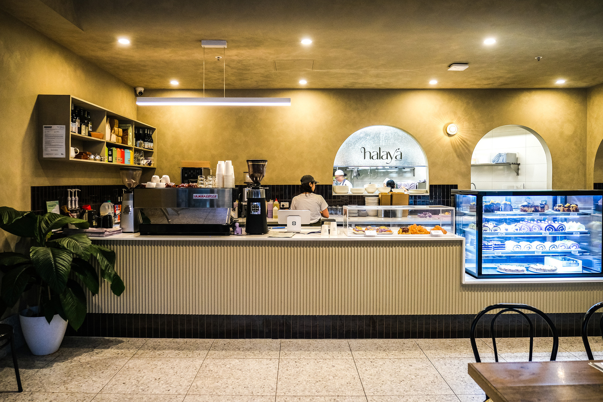 A counter at a cafe with a person making a coffee and a cabinet of desserts
