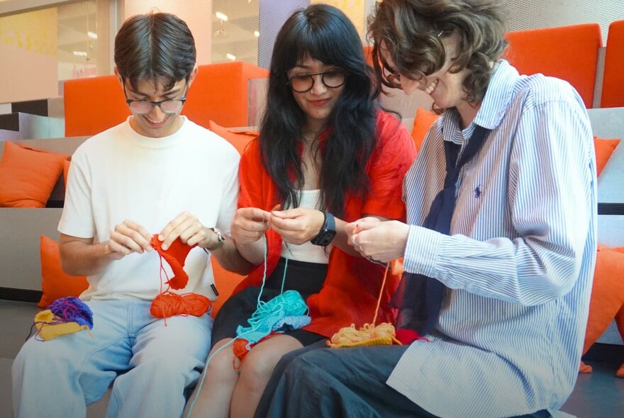 Three people crocheting together on orange chairs.