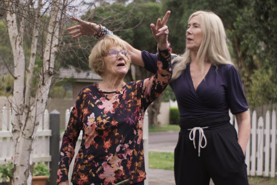 A film still of an older woman pointing to the sky, a younger blonde woman pointing to a birch tree.