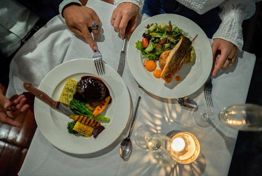 Overhead view of two plates of food, hands holding cutlery, glasses, and a candle on a white linen tablecloth.