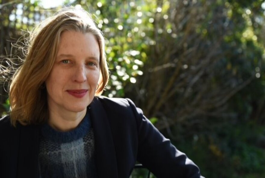 Writer Chloe Hooper, smiling slightly and posed outdoors to the left of the frame, with trees and dappled sunlight behind her. 