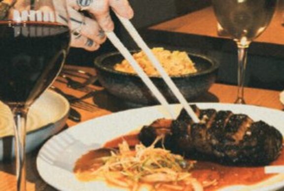 A person's tattooed hand picking up a piece of charred steak with chopsticks.