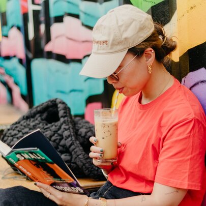 A woman in a beige cap and orange t-shirt sipping an iced coffee while reading a book.