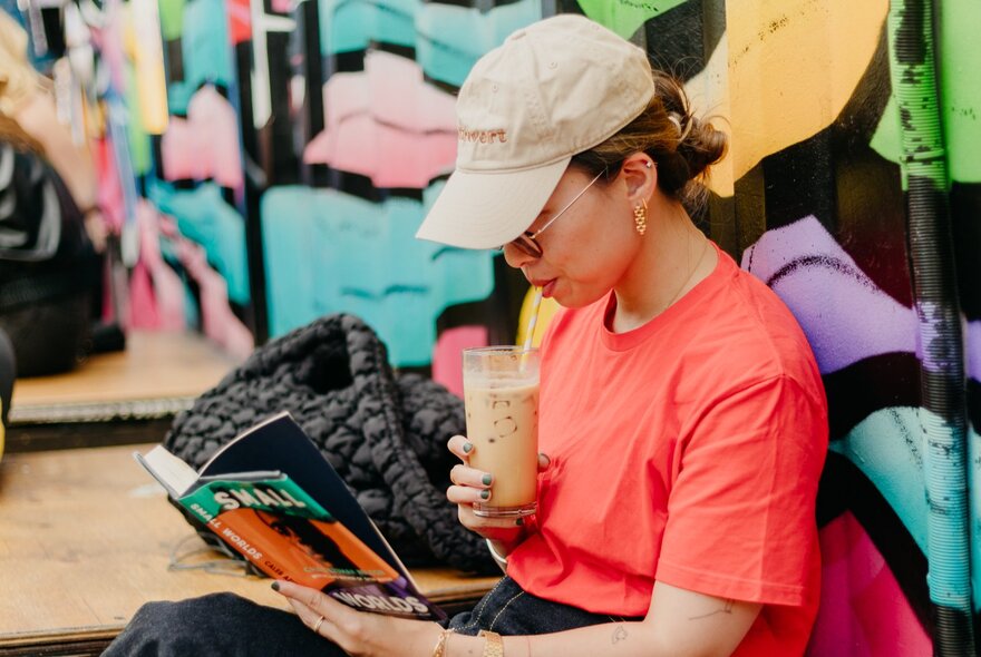 A woman in a beige cap and orange t-shirt sipping an iced coffee while reading a book.