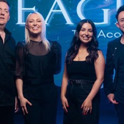 A group of four smiling people standing side-by-side all wearing dark clothing, in front of a blue backdrop, with the word EAGLES partially visible in large white letters.