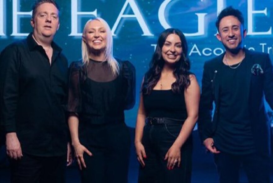 A group of four smiling people standing side-by-side all wearing dark clothing, in front of a blue backdrop, with the word EAGLES partially visible in large white letters.