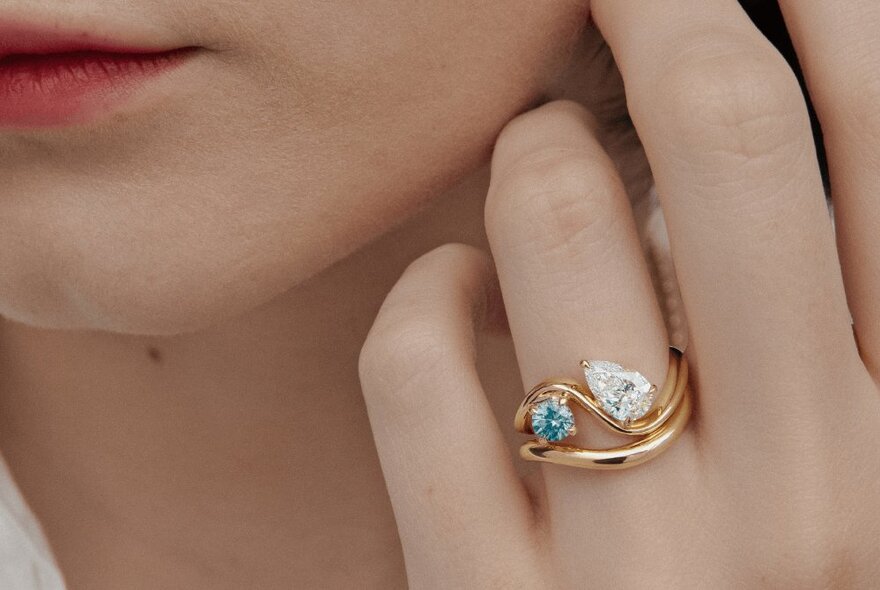 A close-up of a ring with two gemstones and a gold hoop on a woman's hand as she holds it up to her face