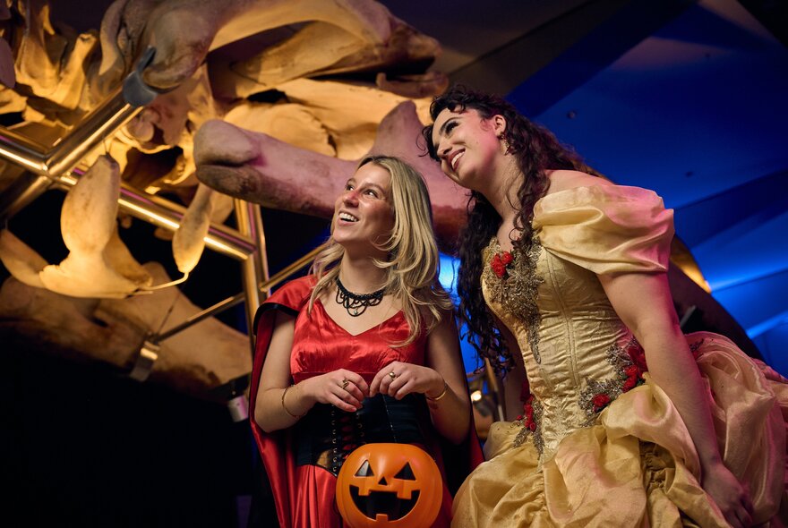 Two women in Halloween costumes looking at a dinosaur fossil in a museum.