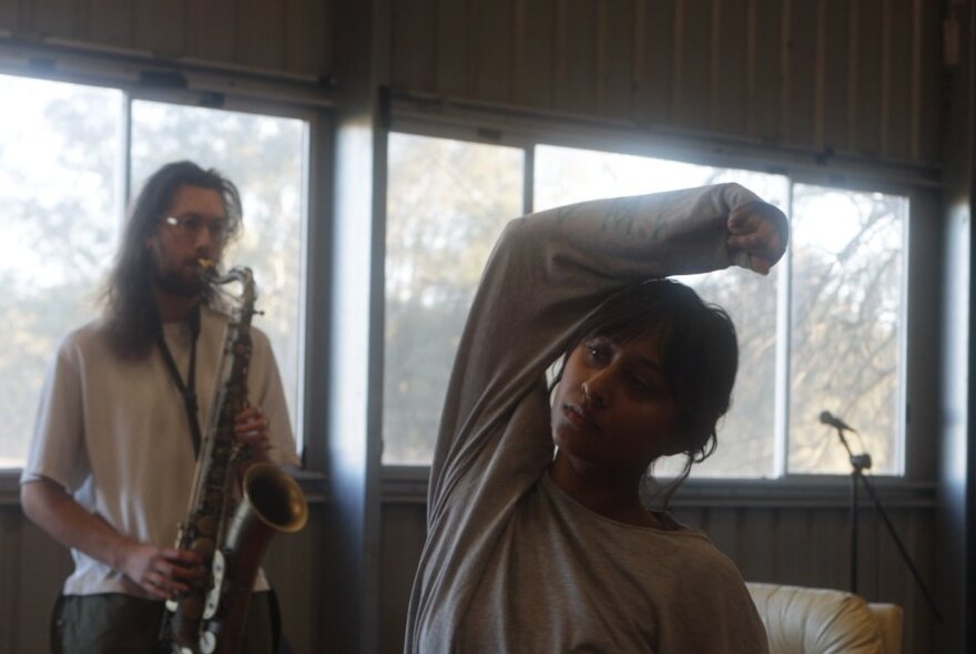 Two people in a room, one of them playing a saxophone and the other person bending their arm over their head.