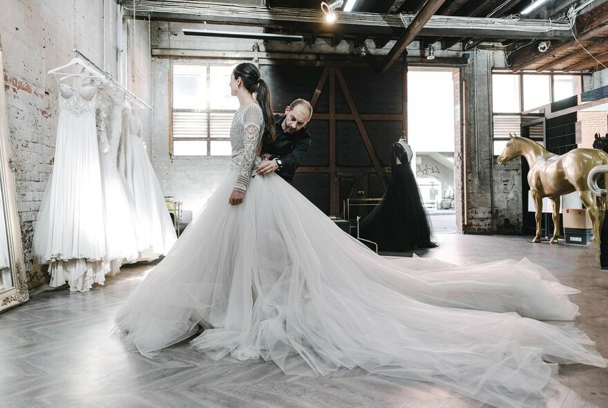 Model being fitted with white dress by a tailor in a warehouse setting.