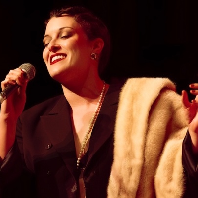 A woman wearing a brown dress with a fur stole draped over her arm, on a darkened stage, singing into a microphone she holds in one hand.