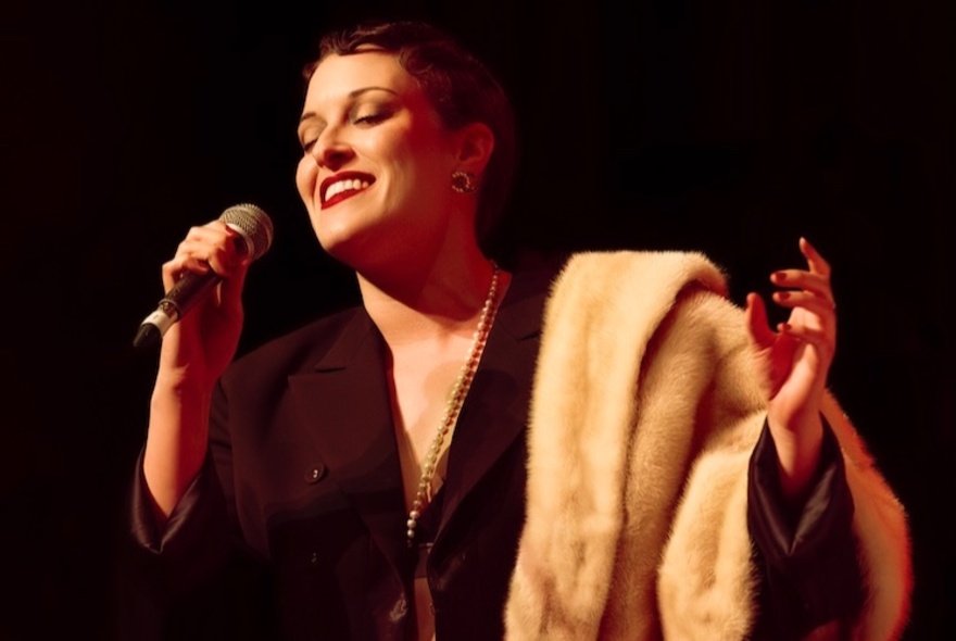 A woman wearing a brown dress with a fur stole draped over her arm, on a darkened stage, singing into a microphone she holds in one hand.