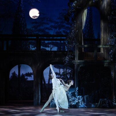 Two classical ballet dancers on stage during a performance of Romeo and Juliet by The Australian Ballet, against a set of a building at night, with a full moon casting a white glow onto the stage.