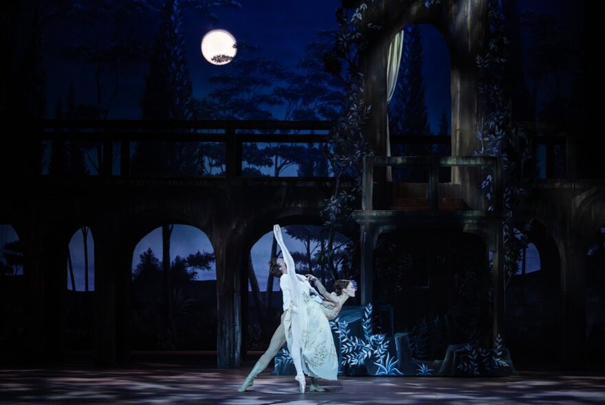 Two classical ballet dancers on stage during a performance of Romeo and Juliet by The Australian Ballet, against a set of a building at night, with a full moon casting a white glow onto the stage.