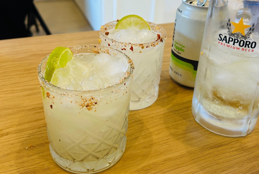 Two glasses of a spicy margarita cocktail garnished with lime wedges alongside a Sapporo beer glass.