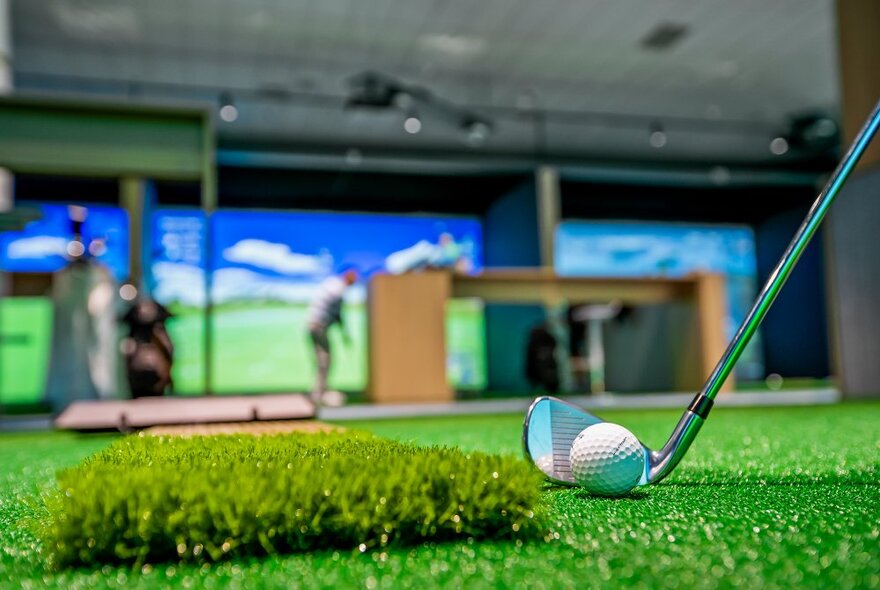 A golf ball and golf club on synthetic turf, in front of individual bays with simulated golf courses on a big screen, indoors.