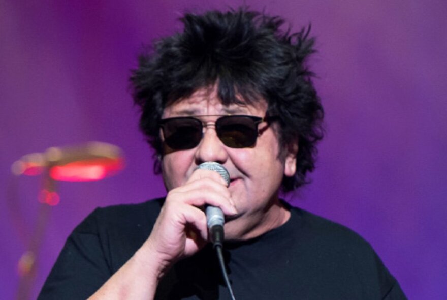 Singer, Richard Clapton, wearing dark sunglasses and singing into a microphone on a stage.