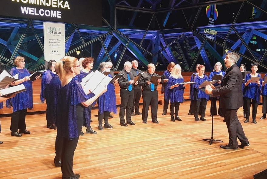 A choir standing and holding sheet music, inside a large auditorium, all faced towards a conductor who stands at the front of the stage.