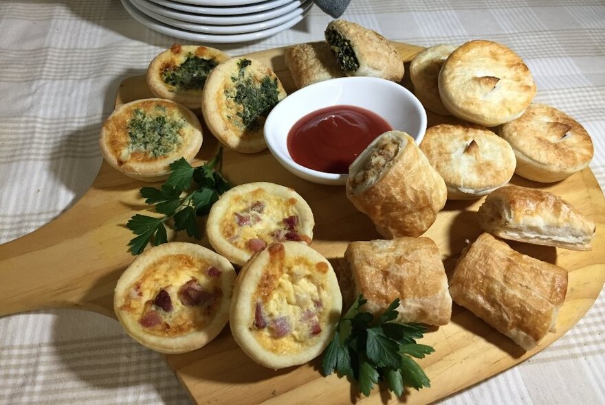 Wooden board with small, savoury baked goods, tomato sauce in small white bowl at centre.