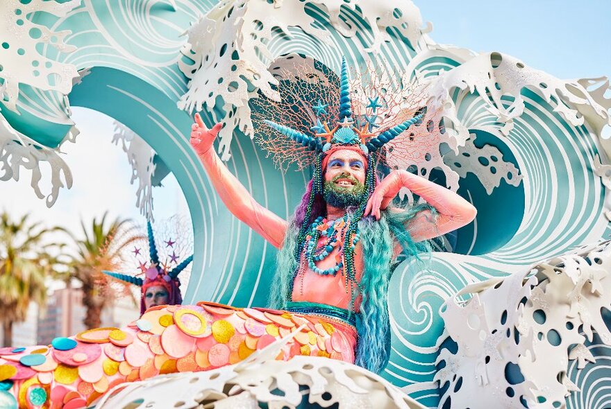 A bearded person dressed as a mermaid and sitting in a float that is designed to look like a large ocean wave.