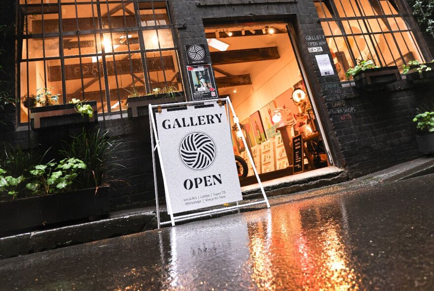 Street entrance to a studio gallery space, with a sandwich board sign out the front, and large windows; evening.