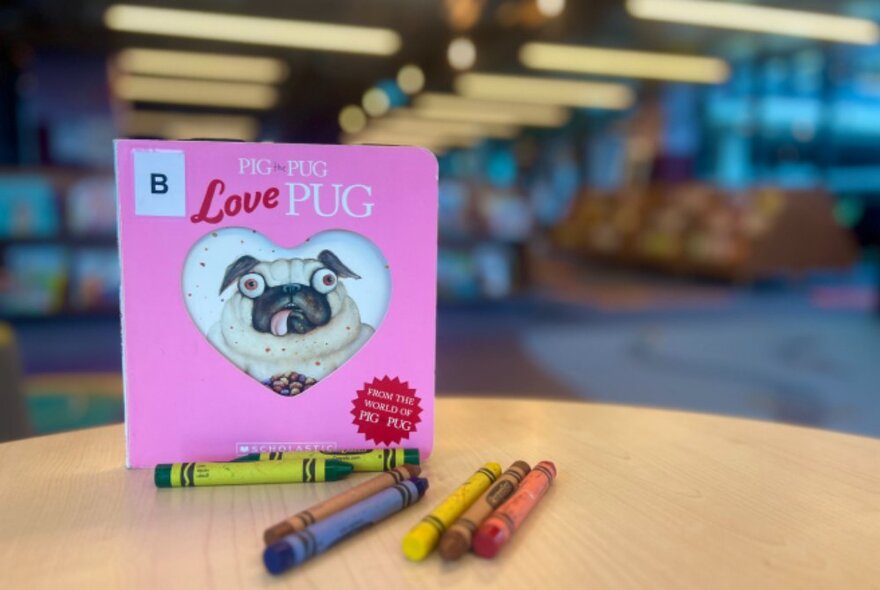 A pink Pig the Pug book on display in a library, with crayons on the table also. 