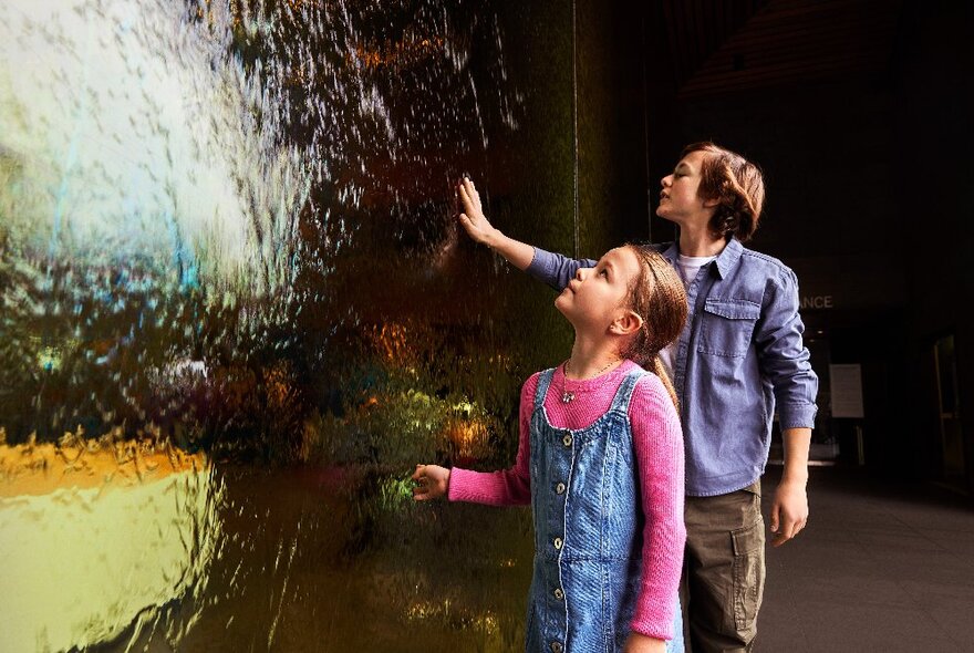 Two children resting their hands on the glass water wall at NGV International.